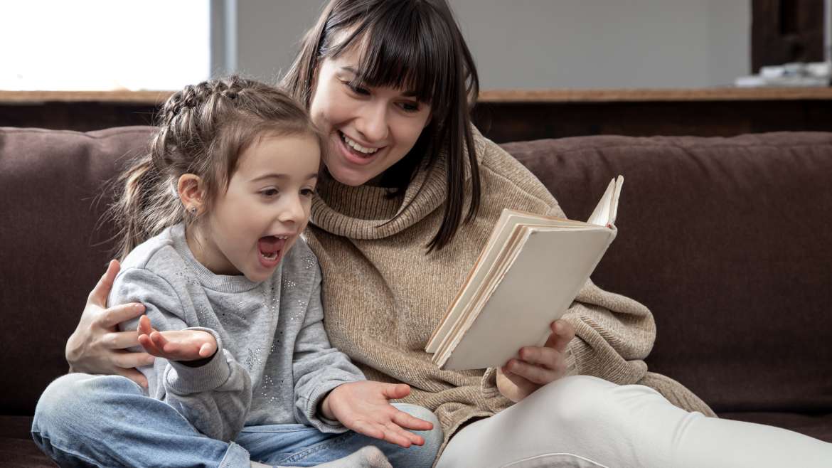 Young mother with her daughter are reading a book at home while sitting on the couch.