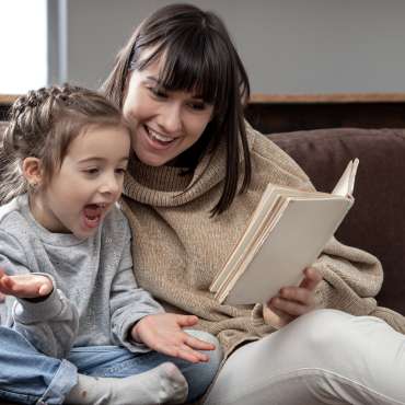 Young mother with her daughter are reading a book at home while sitting on the couch.