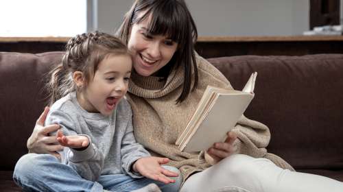 Young mother with her daughter are reading a book at home while sitting on the couch.