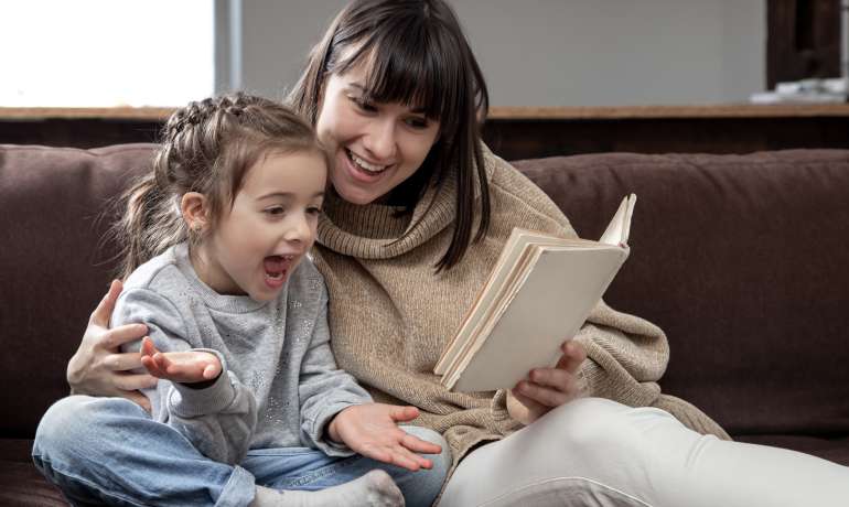 Young mother with her daughter are reading a book at home while sitting on the couch.